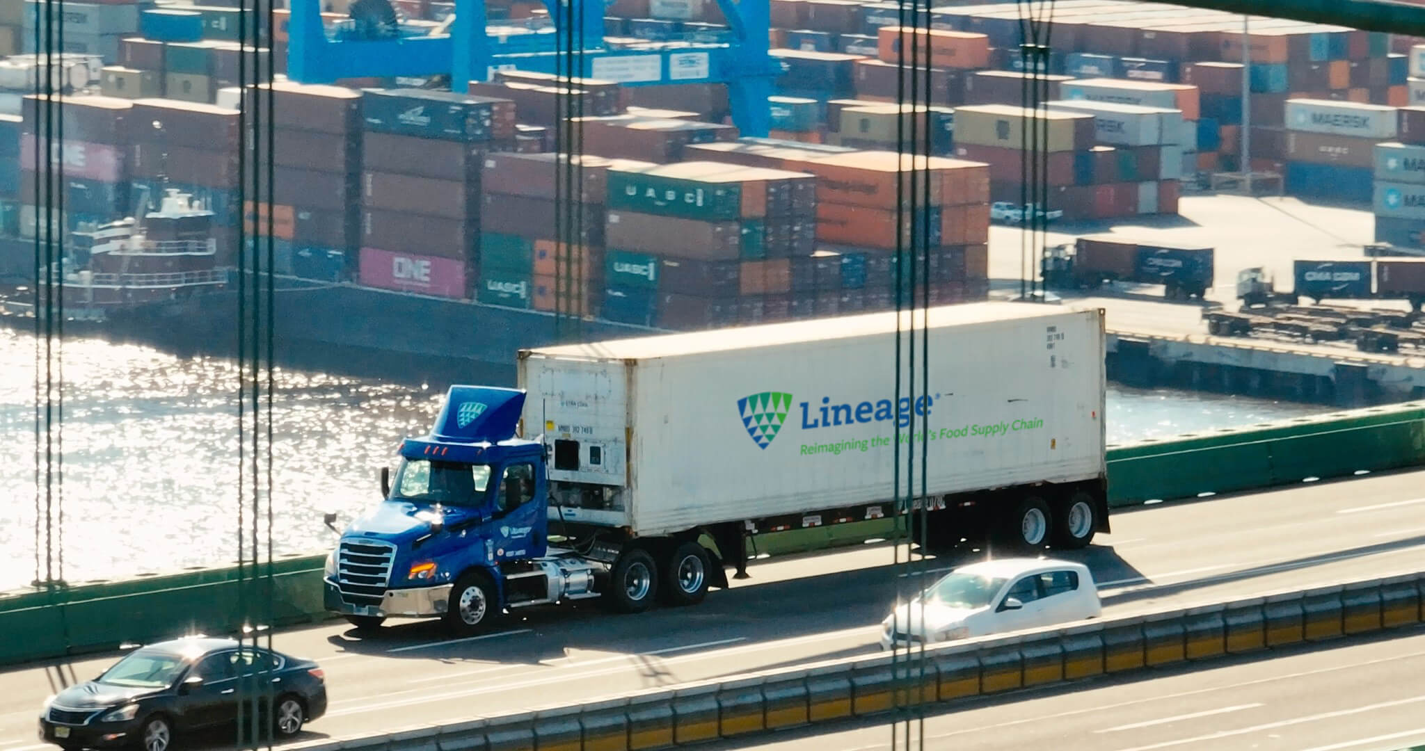 A Lineage truck crossing a bridge with a port full of shipping containers in the background, representing supply chain solutions for navigating tariffs and global trade challenges.