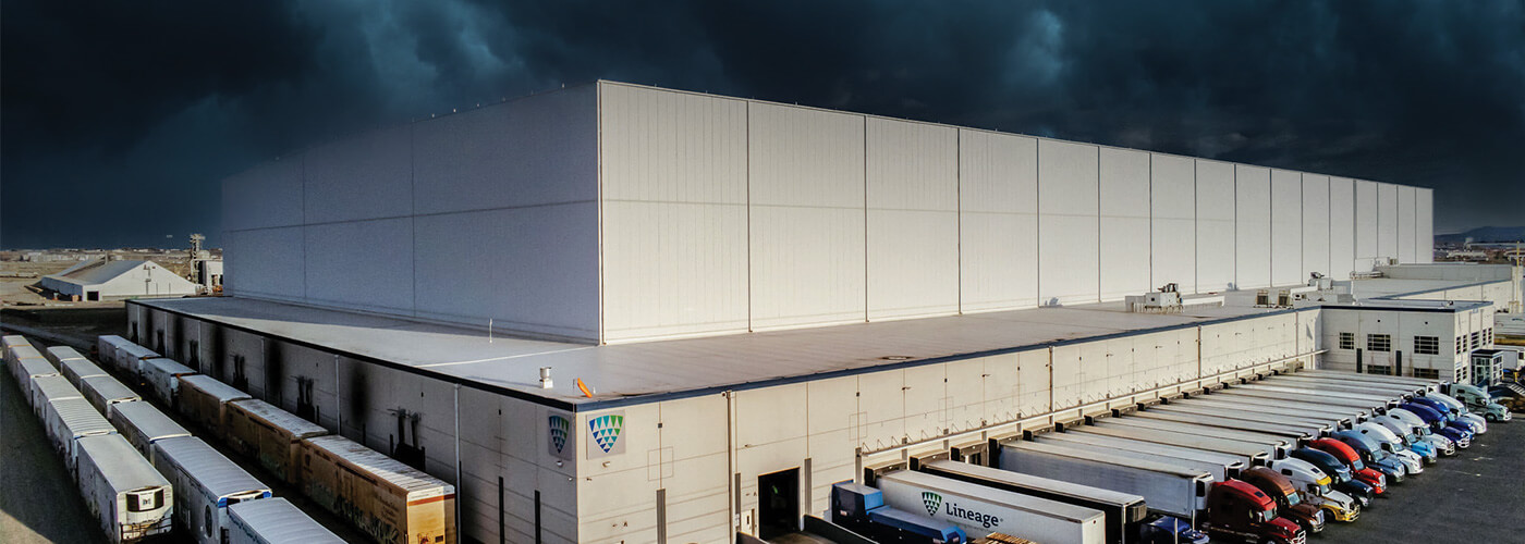A Lineage warehouse under dark storm clouds, with multiple trucks and trailers docked, illustrating reliable cold chain logistics during severe weather conditions.
