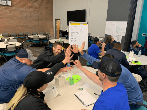 Deelnemers aan het Lineage EDGE-programma nemen deel aan een levendige teambuildingactiviteit binnen, waarbij ze high-fives en enthousiasme uitwisselen in een gezamenlijke werkruimte.