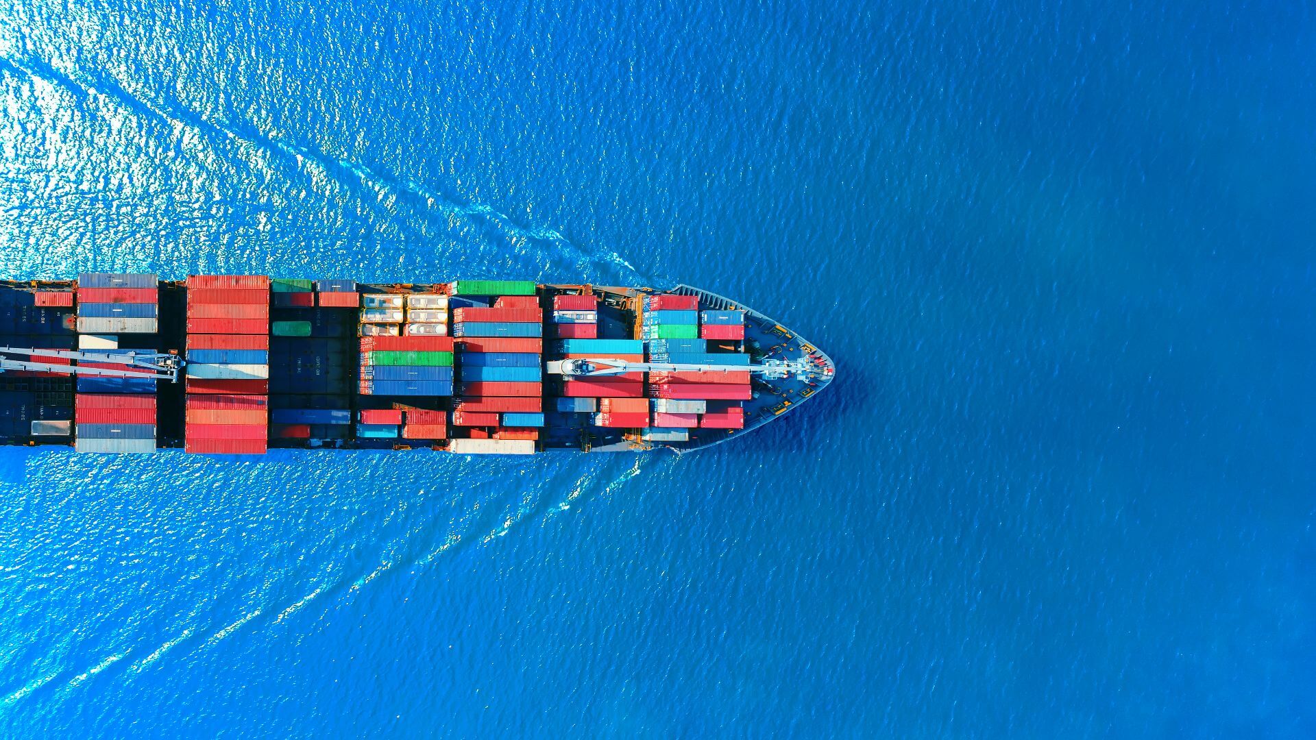 Aerial view of a large cargo ship carrying multicolored shipping containers across a bright blue ocean, symbolizing global trade and port logistics.