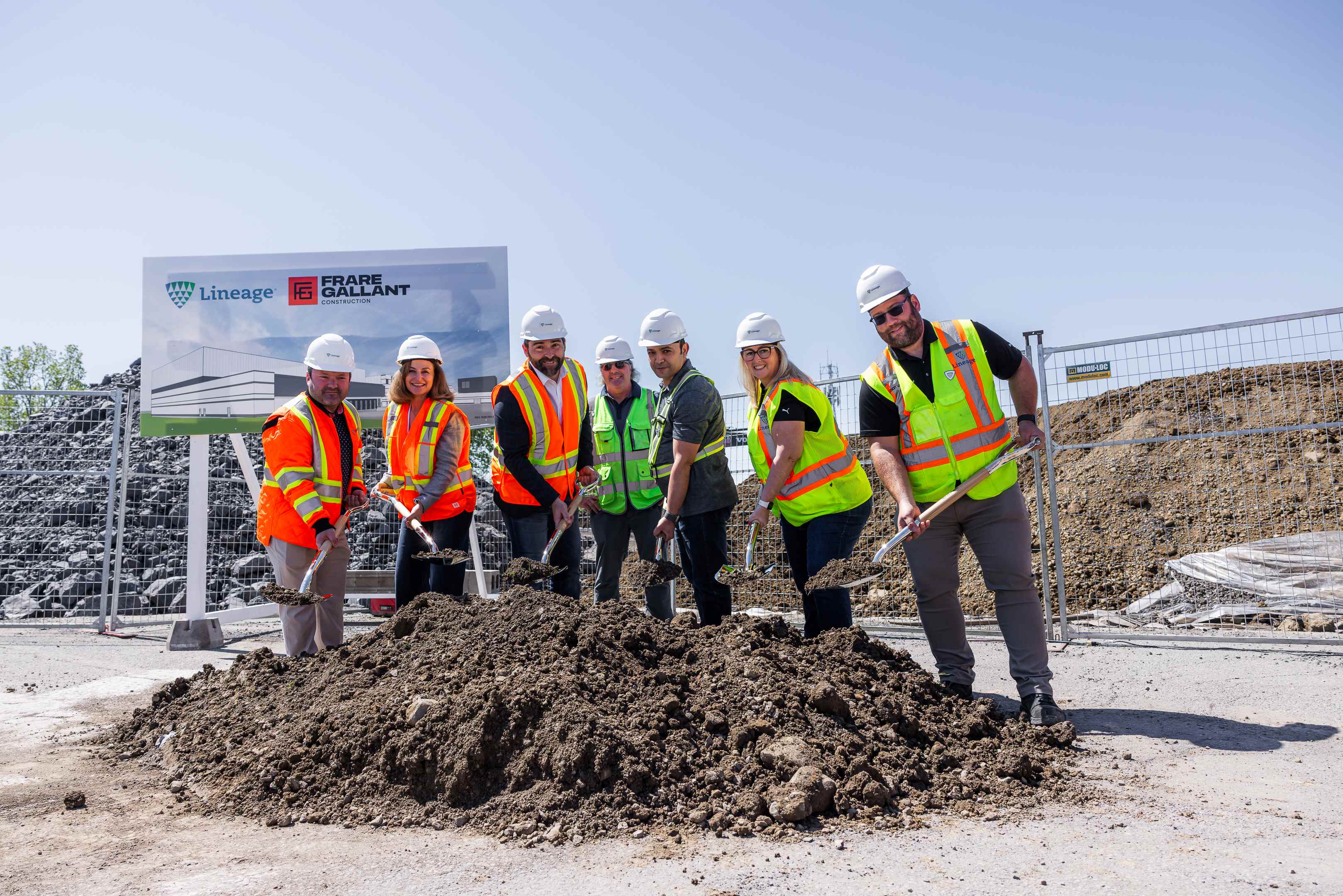 Grupa ludzi w kaskach i kamizelkach odblaskowych stoi z uśmiechem, trzymając łopaty na stosie ziemi na placu budowy.