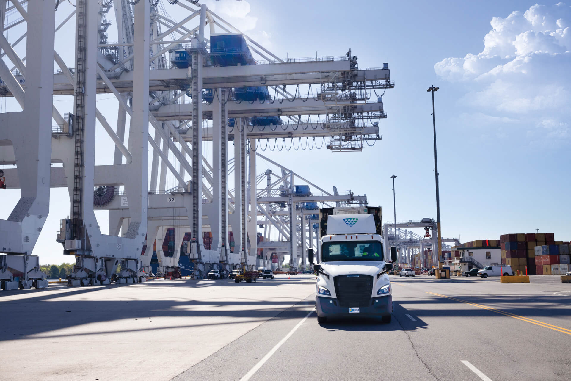 A Lineage truck drives through a busy port terminal, surrounded by large shipping cranes, cargo containers, and port infrastructure under a clear blue sky.