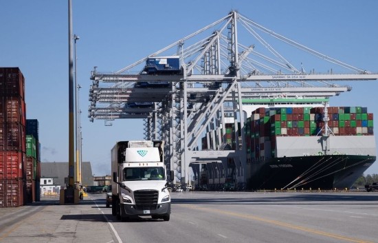 A Lineage refrigerated truck drives through a busy port as cranes unload containers from a cargo vessel, showing the role of drayage in moving imported food quickly to cold storage.