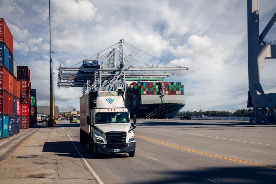 A Lineage-branded refrigerated truck drives through a port terminal with stacked containers and a large cargo ship docked in the background, representing cold chain transport.