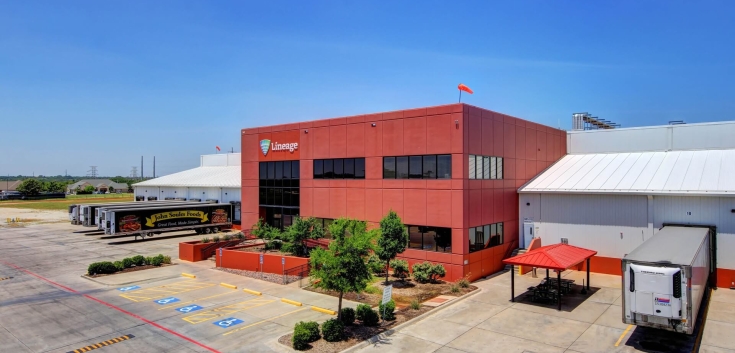 Exterior view of a Lineage cold storage facility in Dallas, Texas, featuring a red office building, adjacent warehouse, parked refrigerated trailers and a clear blue sky.