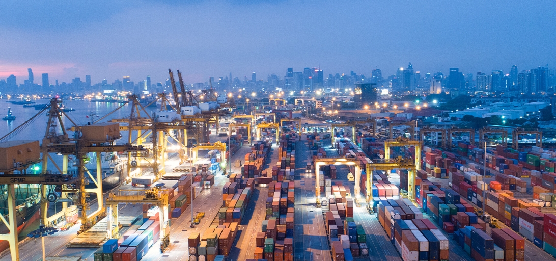 Aerial view of a busy port at dusk with shipping containers, cranes, and city skyline in the background, illustrating global trade and logistics operations.