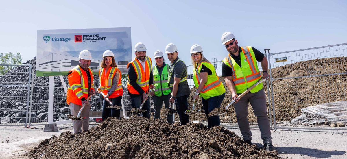 A group of people in hard hats and high vis safety vests stand smiling while holding shovels in a pile of dirt at a construction site