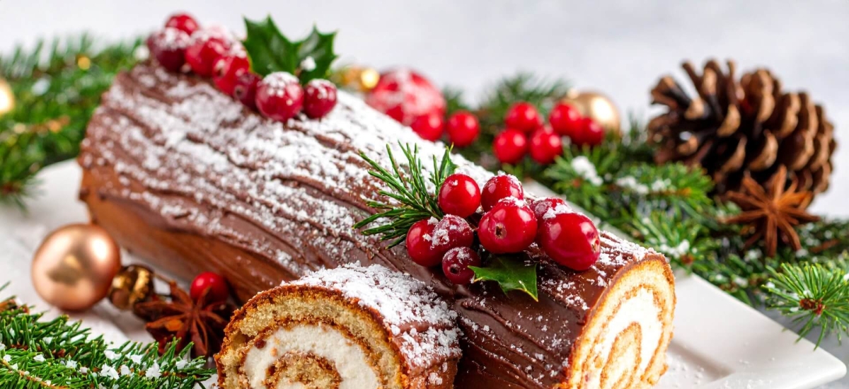 A chocolate yule log cake decorated with cranberries, holly and powdered sugar on a holiday-themed plate.
