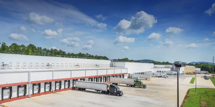 A view of Lineage's cold storage warehouse in Attalla, Alabama, showcasing multiple loading docks with semi-trucks parked for deliveries. The large white facility is surrounded by an expansive concrete lot and green landscaping under a bright blue sky with scattered clouds.