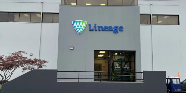 Der Haupteingang der Lineage Niederlassung in Lineage mit dem Firmenlogo und einer hell erleuchteten, modernen Fassade vor bewölktem Himmel.