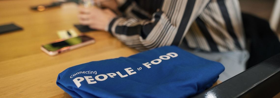 Hay una camisa azul con texto blanco que dice “Conectando personas con alimentos” doblada sobre una mesa