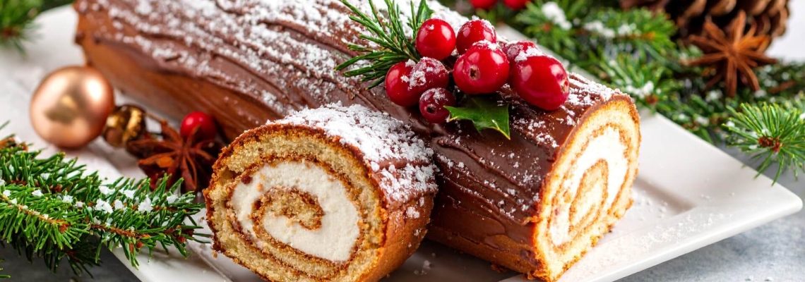 A chocolate yule log cake decorated with cranberries, holly and powdered sugar on a holiday-themed plate.