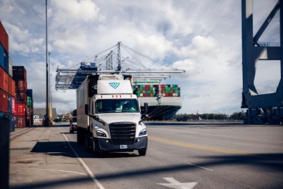 Temperature-controlled truck hauling a container at a busy seaport with cargo ship and cranes in the background, illustrating U.S. food export logistics and drayage operations.