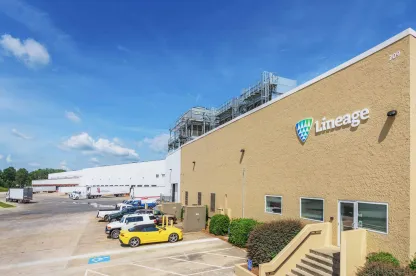 A Lineage cold storage warehouse in Attalla, Alabama, with the company's logo displayed on the beige exterior. The image shows a row of parked vehicles, including a yellow sports car, a loading dock area with semi-trucks, and the facility's infrastructure under a clear blue sky.