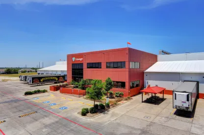 Exterior view of a Lineage cold storage facility in Dallas, Texas, featuring a red office building, adjacent warehouse, parked refrigerated trailers and a clear blue sky.